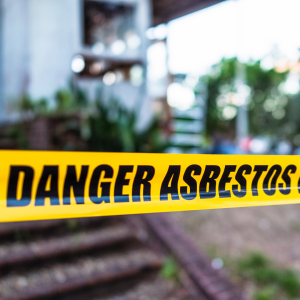 Close-up of yellow 'Danger Asbestos' tape in front of a blurred background with a building and greenery, signaling an asbestos hazard on site