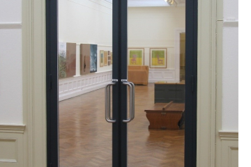 Glass fire doors with a black frame installed in a gallery setting, providing clear visibility while maintaining fire safety. The doors feature silver handles and overhead door closers. A certification mark in the corner indicates compliance with UKCA and EN14449 standards, ensuring fire resistance and safety compliance.