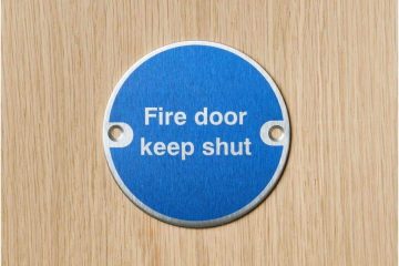 Close-up of a circular blue fire safety sign mounted on a wooden fire door. The sign reads 'Fire door keep shut' in white text, emphasizing the importance of keeping the door closed to maintain fire safety and compliance with regulations.